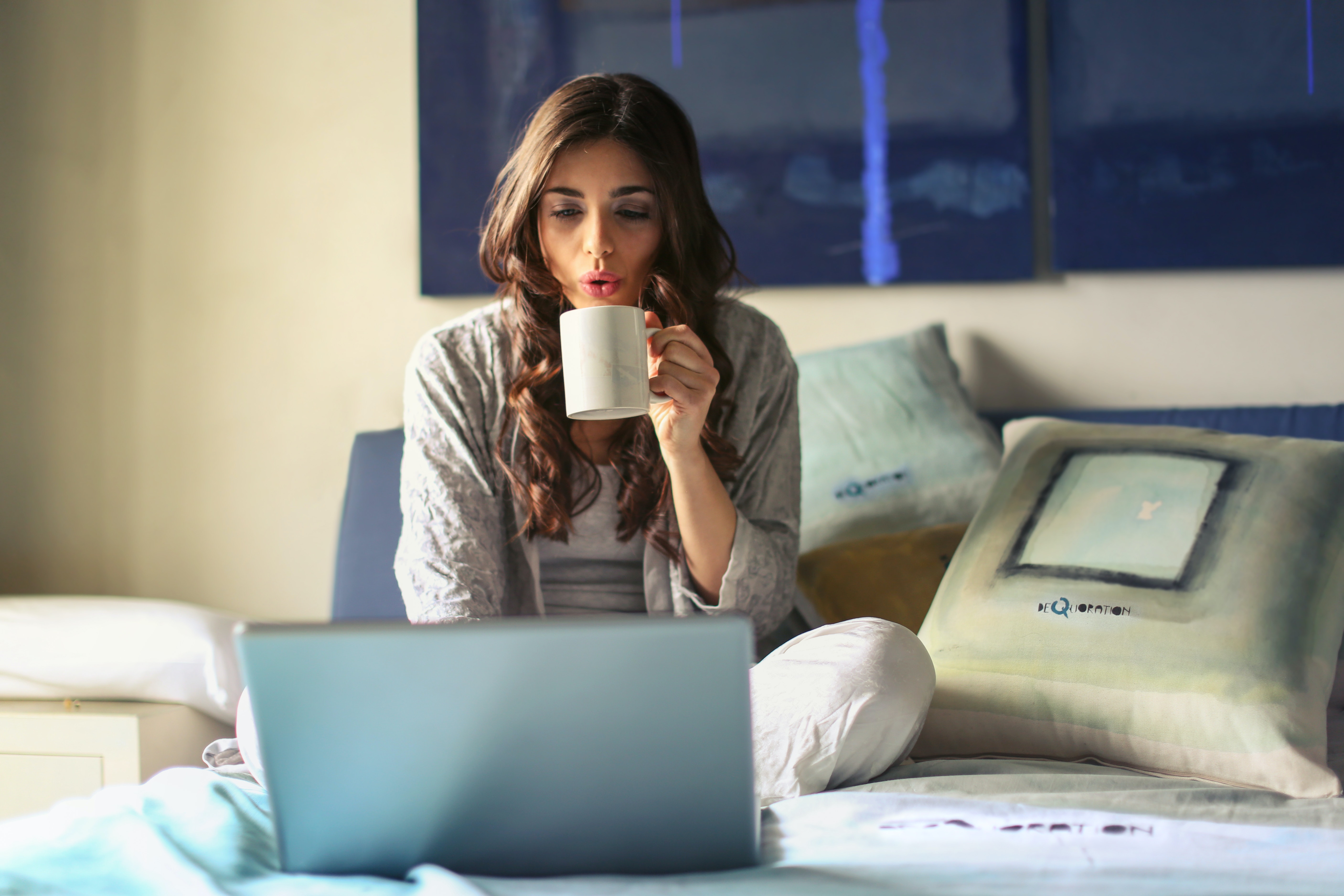 woman sitting on bed, sipping from a mug, on a laptop